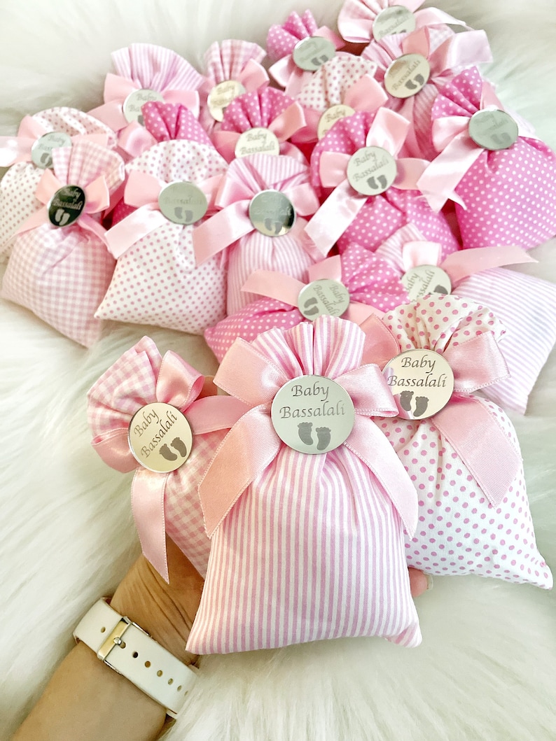 May include: Pink and white fabric gift bags with a silver tag that says "Baby Bassalali" and a pink ribbon. The bags are decorated with polka dots and stripes.