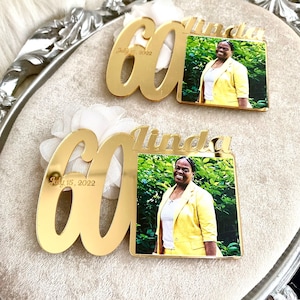May include: Two gold mirror acrylic plaques with the number 60 and the name Linda. The plaques have a photo of a woman in a yellow blazer. The text "July 15, 2022" is engraved on the plaques.