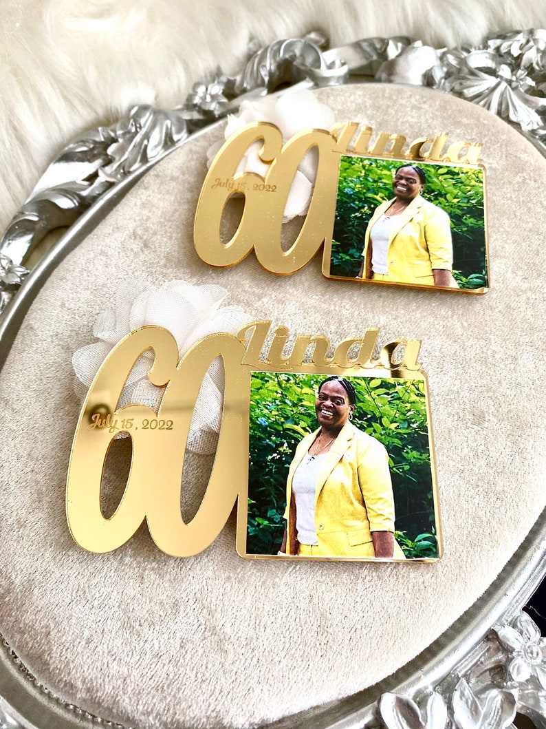 May include: Two gold mirror acrylic plaques with the number 60 and the name Linda. The plaques have a photo of a woman in a yellow blazer. The text "July 15, 2022" is engraved on the plaques.