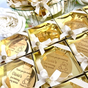 May include: Gold favor boxes with a round mirror inside. Each mirror has a personalized message that says "Thank you for making Olivia's first birthday very special" with a teddy bear graphic and the date March 30, 2022.