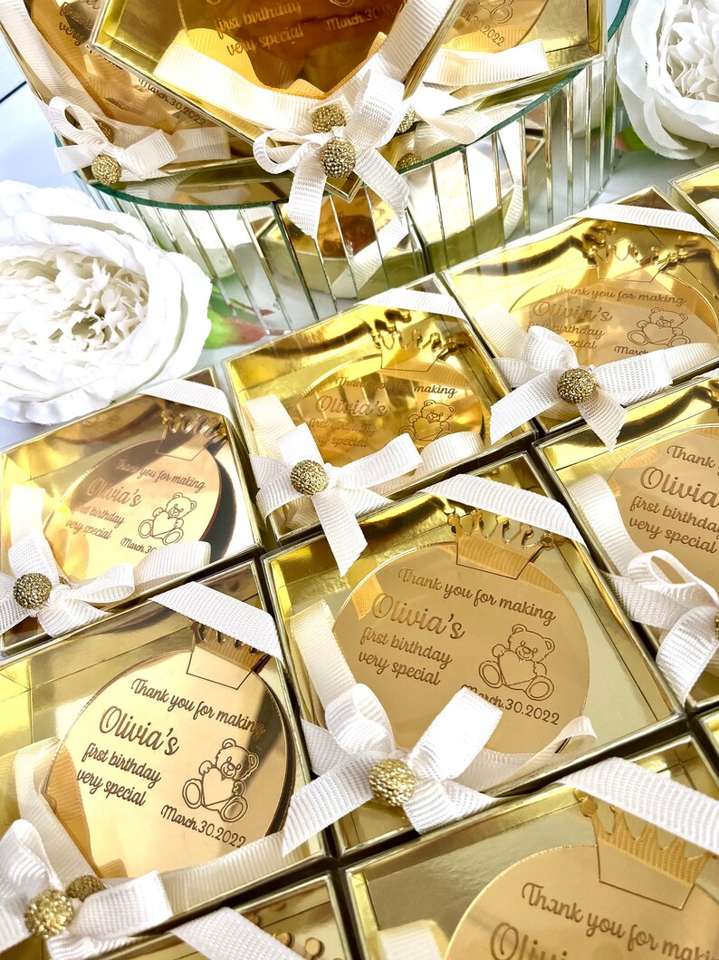 May include: Gold favor boxes with a round mirror inside. Each mirror has a personalized message that says "Thank you for making Olivia's first birthday very special" with a teddy bear graphic and the date March 30, 2022.