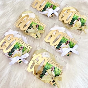 May include: Six gold-colored, personalized 60th birthday ornaments with a photo of a woman in each. Each ornament is in a clear plastic box with a white ribbon and a gold bow. The ornaments are labeled with the name "Linda" and the date "July 15, 2022".