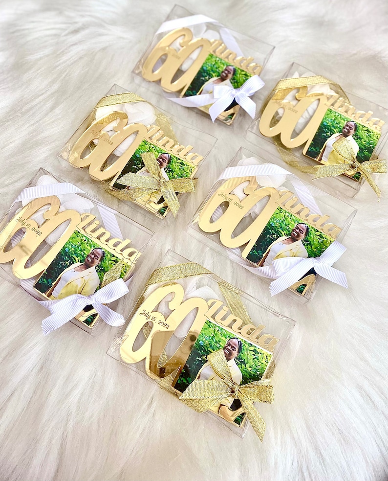 May include: Six gold-colored, personalized 60th birthday ornaments with a photo of a woman in each. Each ornament is in a clear plastic box with a white ribbon and a gold bow. The ornaments are labeled with the name "Linda" and the date "July 15, 2022".