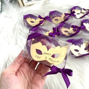 Puede incluir: Espejo dorado en forma de m&aacute;scara en una caja de pl&aacute;stico transparente con una cinta y una pluma moradas. El espejo tiene el texto "Fifty & Fabulous" y "Jeane's" grabado.