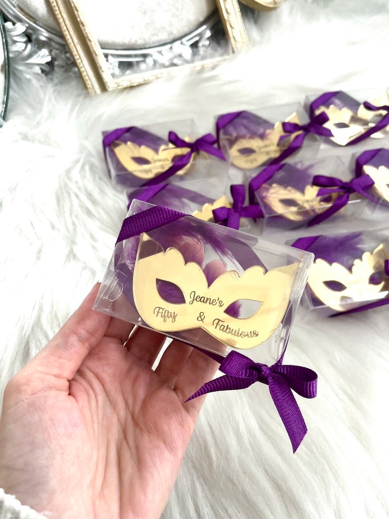 Puede incluir: Espejo dorado en forma de m&aacute;scara en una caja de pl&aacute;stico transparente con una cinta y una pluma moradas. El espejo tiene el texto "Fifty & Fabulous" y "Jeane's" grabado.