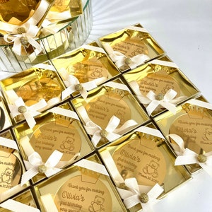 May include: Gold favor boxes with a round, gold mirror with the text "Thank you for making Olivia's first birthday very special March 30, 2022" and a small teddy bear graphic.
