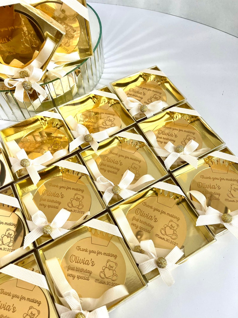 May include: Gold favor boxes with a round, gold mirror with the text "Thank you for making Olivia's first birthday very special March 30, 2022" and a small teddy bear graphic.