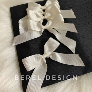 May include: A black fabric pouch with white satin ribbon bows tied on the front. The pouch has the text "BEREL DESIGN" printed on the front.