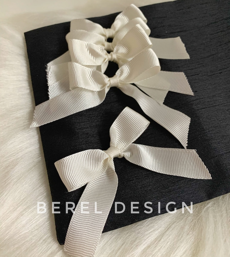May include: A black fabric pouch with white satin ribbon bows tied on the front. The pouch has the text "BEREL DESIGN" printed on the front.