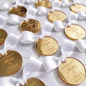 May include: A collection of circular gold-coloured acrylic tags, each featuring a white satin ribbon bow. The tags are engraved with the text The Wedding Of Matta & Kara 3rd February 2025, arranged in a repeating pattern across the frame.