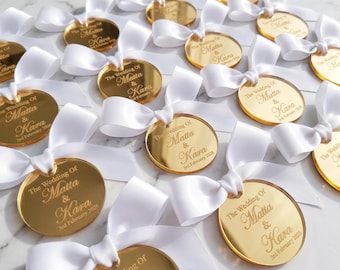 May include: A collection of circular gold-colored acrylic tags, each featuring a white satin ribbon bow. The tags are engraved with the text The Wedding Of Matta & Kara 3rd February 2025, arranged in a repeating pattern across the frame.