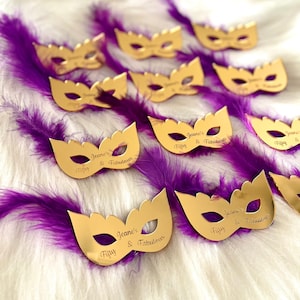 Puede incluir: M&aacute;scaras doradas de carnaval con el texto "Jeane's Fifty & Fabulous" grabado en ellas. Las m&aacute;scaras est&aacute;n colocadas sobre una superficie blanca y esponjosa con plumas moradas.