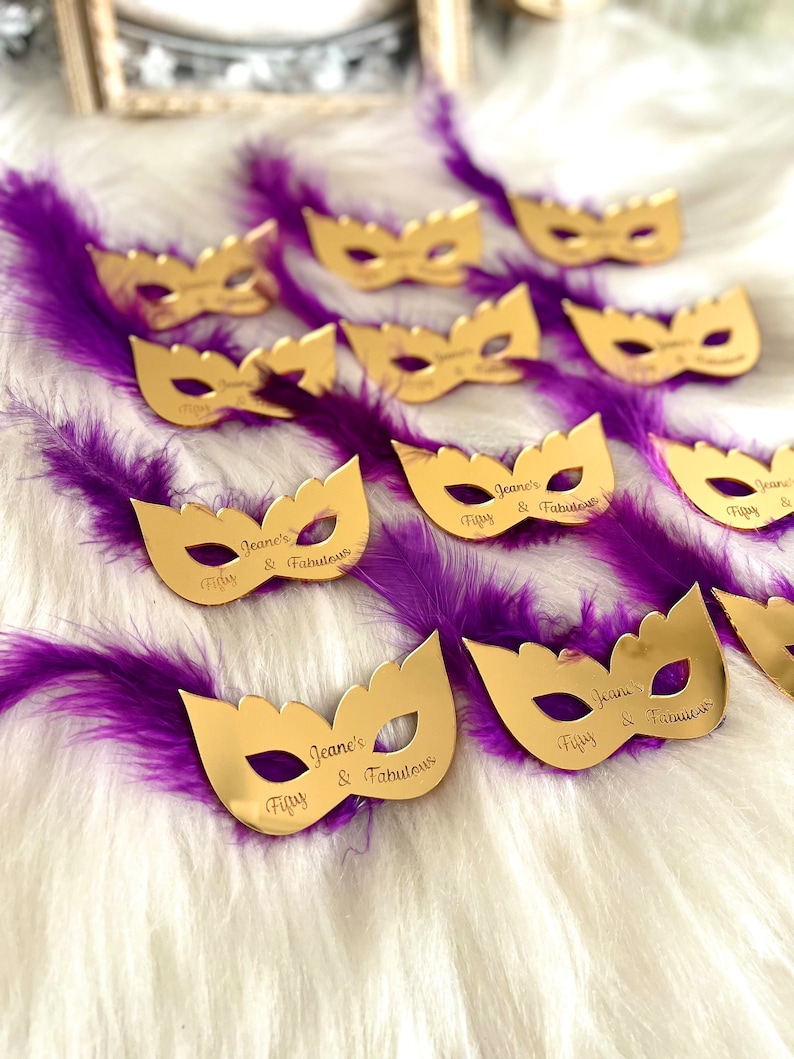 Puede incluir: M&aacute;scaras doradas de carnaval con el texto "Jeane's Fifty & Fabulous" grabado en ellas. Las m&aacute;scaras est&aacute;n colocadas sobre una superficie blanca y esponjosa con plumas moradas.