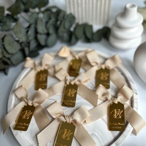 May include: Gold favor tags with a white satin ribbon bow. Each tag features a gold letter "E" and the text "Dr. Evelyn Muncha 02/10/2023".