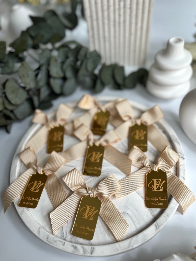 May include: Gold favor tags with a white satin ribbon bow. Each tag features a gold letter "E" and the text "Dr. Evelyn Muncha 02/10/2023".