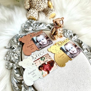 May include: Three silver, gold, and rose gold bear-shaped photo frames with the name "Oliver" engraved on them. Each frame has a photo of a baby and the date "September 24th, 2022".