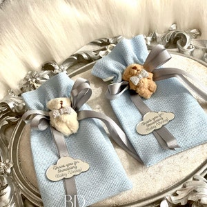 Puede incluir: Dos bolsas de regalo azul claro con un peque&ntilde;o osito de peluche con una pajarita atada a cada una. Las bolsas est&aacute;n atadas con una cinta plateada y tienen una etiqueta plateada con el texto "&iexcl;Gracias por celebrar a Baby Vinny!"