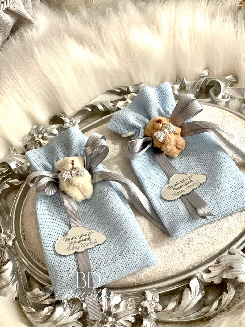 Puede incluir: Dos bolsas de regalo azul claro con un peque&ntilde;o osito de peluche con una pajarita atada a cada una. Las bolsas est&aacute;n atadas con una cinta plateada y tienen una etiqueta plateada con el texto "&iexcl;Gracias por celebrar a Baby Vinny!"