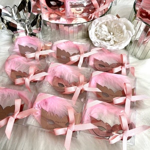 May include: Pink and gold favour boxes with a pink feather and a gold mask inside. The boxes are tied with a pink ribbon and have a personalised tag that says "Sienna's" and "May 6, 2023".