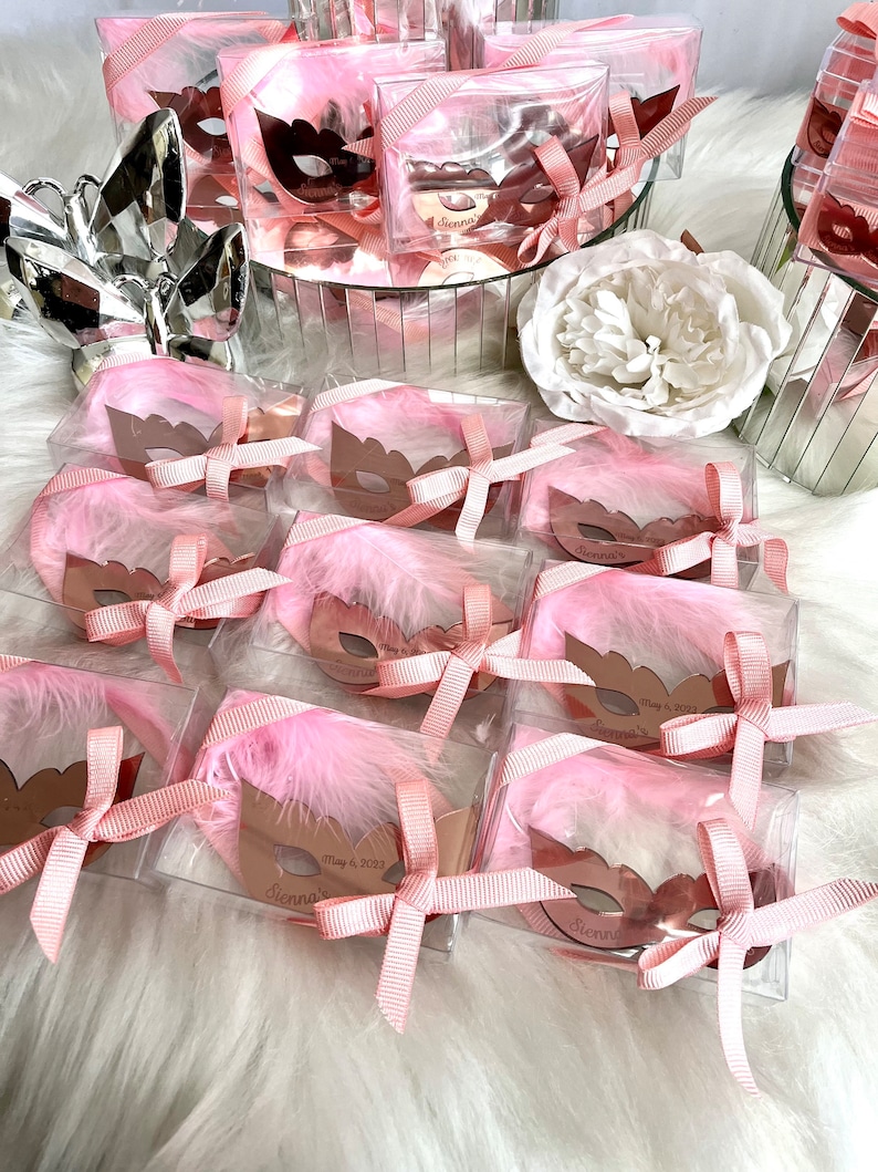 May include: Pink and gold favour boxes with a pink feather and a gold mask inside. The boxes are tied with a pink ribbon and have a personalised tag that says "Sienna's" and "May 6, 2023".