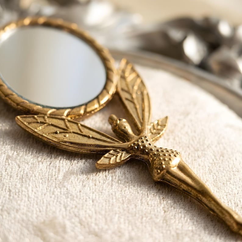 May include: A gold-toned handheld mirror featuring a circular glass surface framed by a textured border. The handle is shaped like a winged figure with detailed leaf-like wings and a textured body, extending into a tapered, smooth handle base.