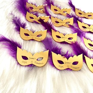 Puede incluir: M&aacute;scaras doradas de carnaval con plumas moradas. Cada m&aacute;scara tiene el texto "Jeane's Fifty & Fabulous" grabado.