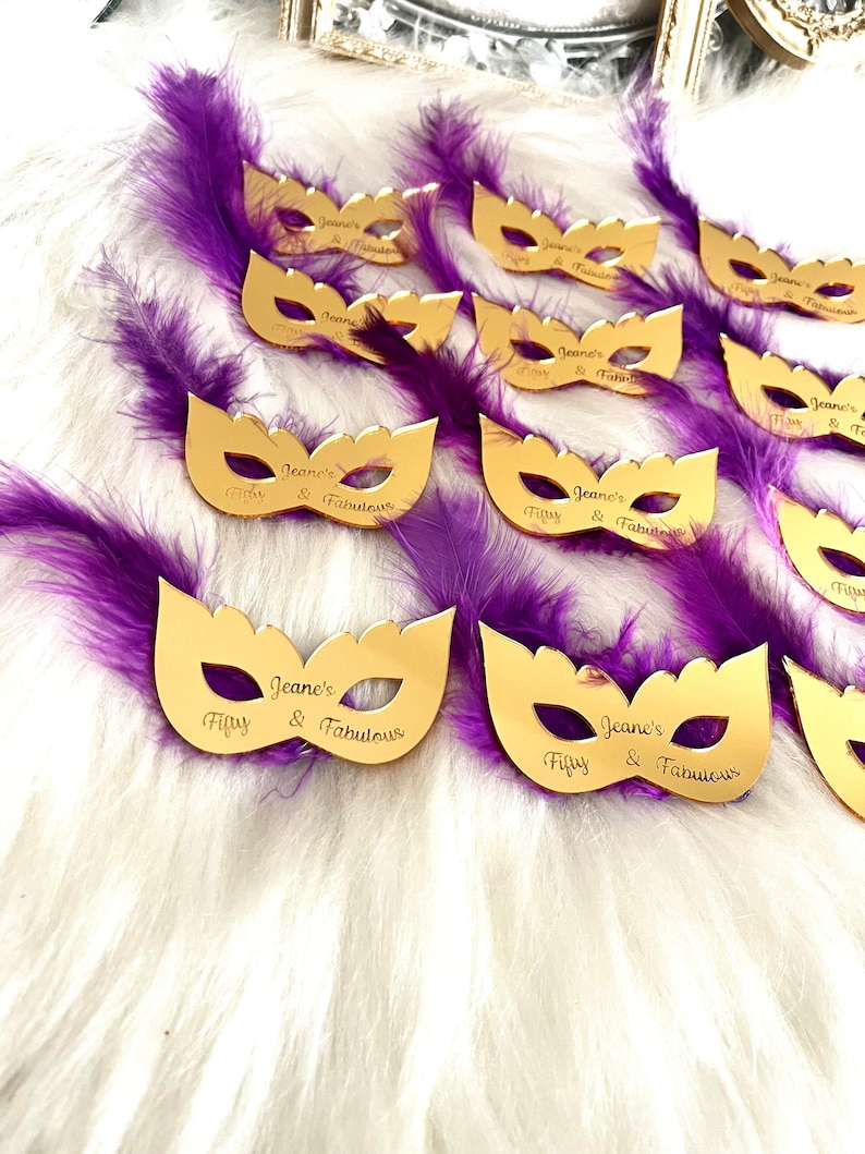 Puede incluir: M&aacute;scaras doradas de carnaval con plumas moradas. Cada m&aacute;scara tiene el texto "Jeane's Fifty & Fabulous" grabado.