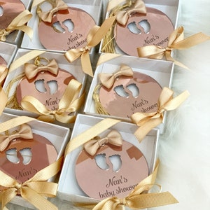 Op de afbeelding: Een set kleine, ronde, roze gouden spiegelversieringen met een uitsnijding van babyvoetjes en de tekst "Nan's baby shower" in een witte doos met een gouden lint.