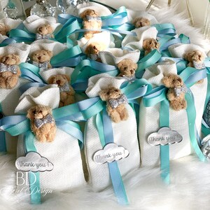 May include: White favor bags with a small teddy bear attached to each bag. The bags are tied with a blue and turquoise ribbon and have a silver tag that says "Thank you!"