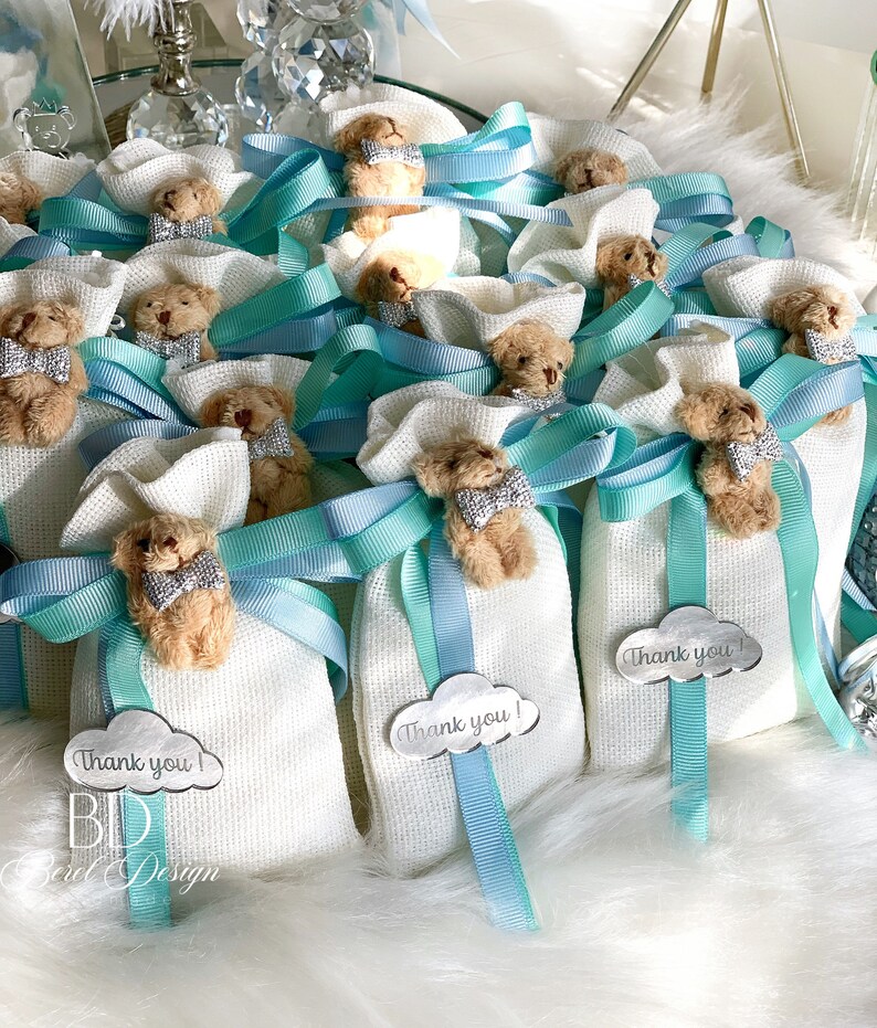 May include: White favor bags with a small teddy bear attached to each bag. The bags are tied with a blue and turquoise ribbon and have a silver tag that says "Thank you!"