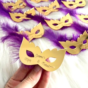 Puede incluir: M&aacute;scara de espejo dorada con plumas moradas y el texto "Jeane's Fifty & Fabulous" grabado en la m&aacute;scara.