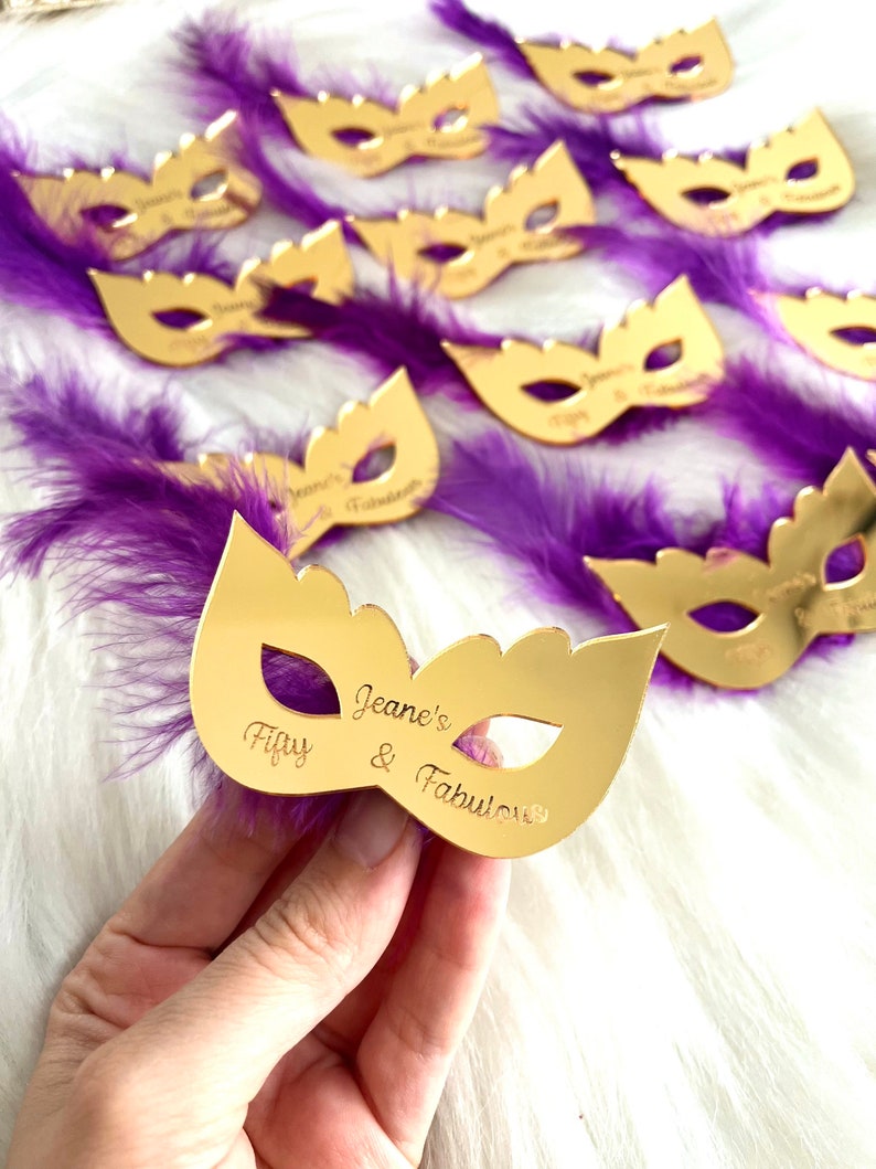 Puede incluir: M&aacute;scara de espejo dorada con plumas moradas y el texto "Jeane's Fifty & Fabulous" grabado en la m&aacute;scara.