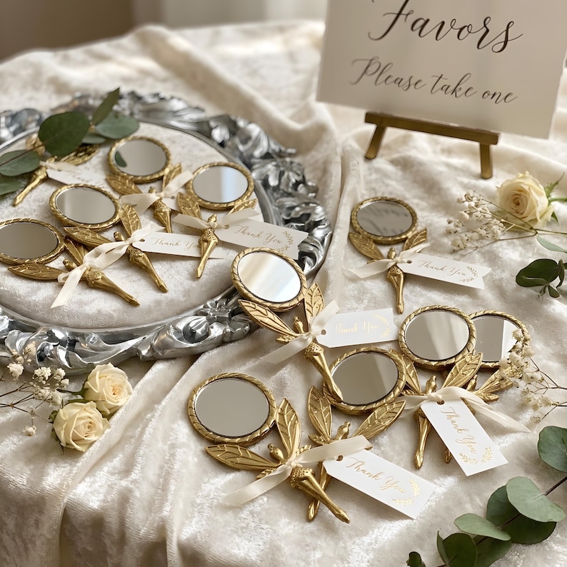 May include: A collection of gold-toned handheld mirrors with dragonfly-shaped handles, each tied with a cream-colored ribbon and a small white tag printed with the words Thank You. The mirrors are arranged on a silver tray and a white fabric surface.