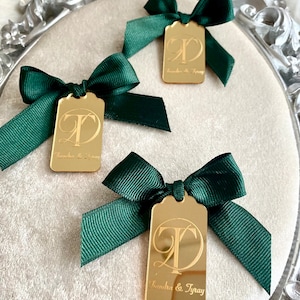 May include: Three gold mirror tags with green satin bows. Each tag features a calligraphic letter "T" and the text "Sandra & Tyray".