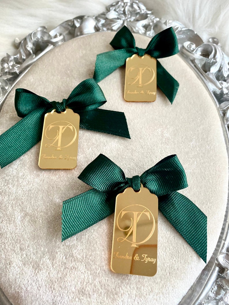 May include: Three gold mirror tags with green satin bows. Each tag features a calligraphic letter "T" and the text "Sandra & Tyray".