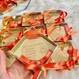 May include: Gold favor boxes with orange ribbon and a small round mirror with a personalized message. The message reads "Thank you for making Mathias' 1st birthday very special. Feb. 22, 2022".