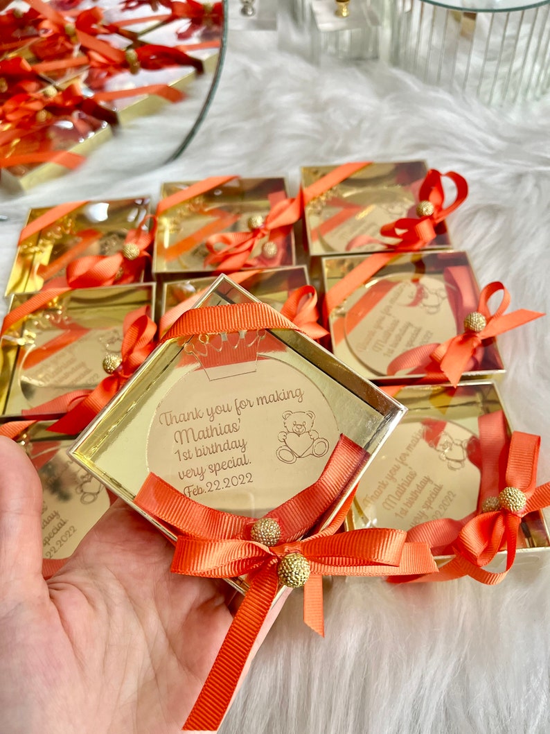 May include: Gold favor boxes with orange ribbon and a small round mirror with a personalized message. The message reads "Thank you for making Mathias' 1st birthday very special. Feb. 22, 2022".