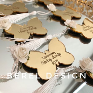 Puede incluir: Favores de espejo en forma de mariposa dorada con borlas blancas y una flor blanca. El espejo tiene el texto "Welcoming Baby Aoufe 2019" grabado.