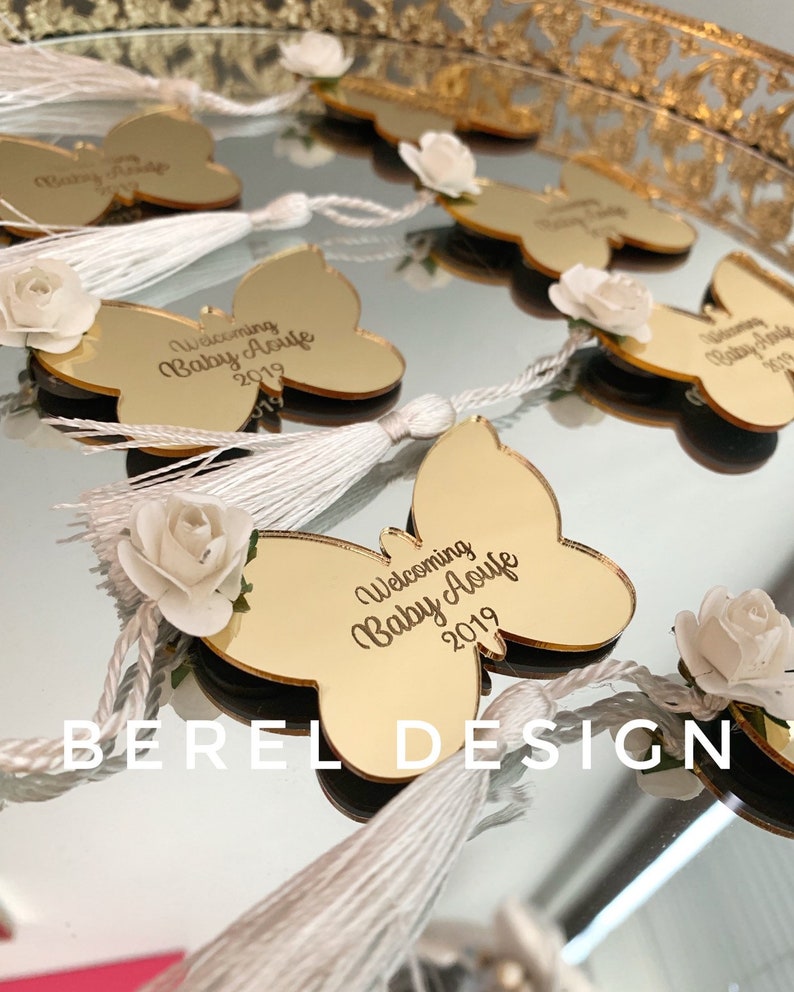 Puede incluir: Favores de espejo en forma de mariposa dorada con borlas blancas y una flor blanca. El espejo tiene el texto "Welcoming Baby Aoufe 2019" grabado.