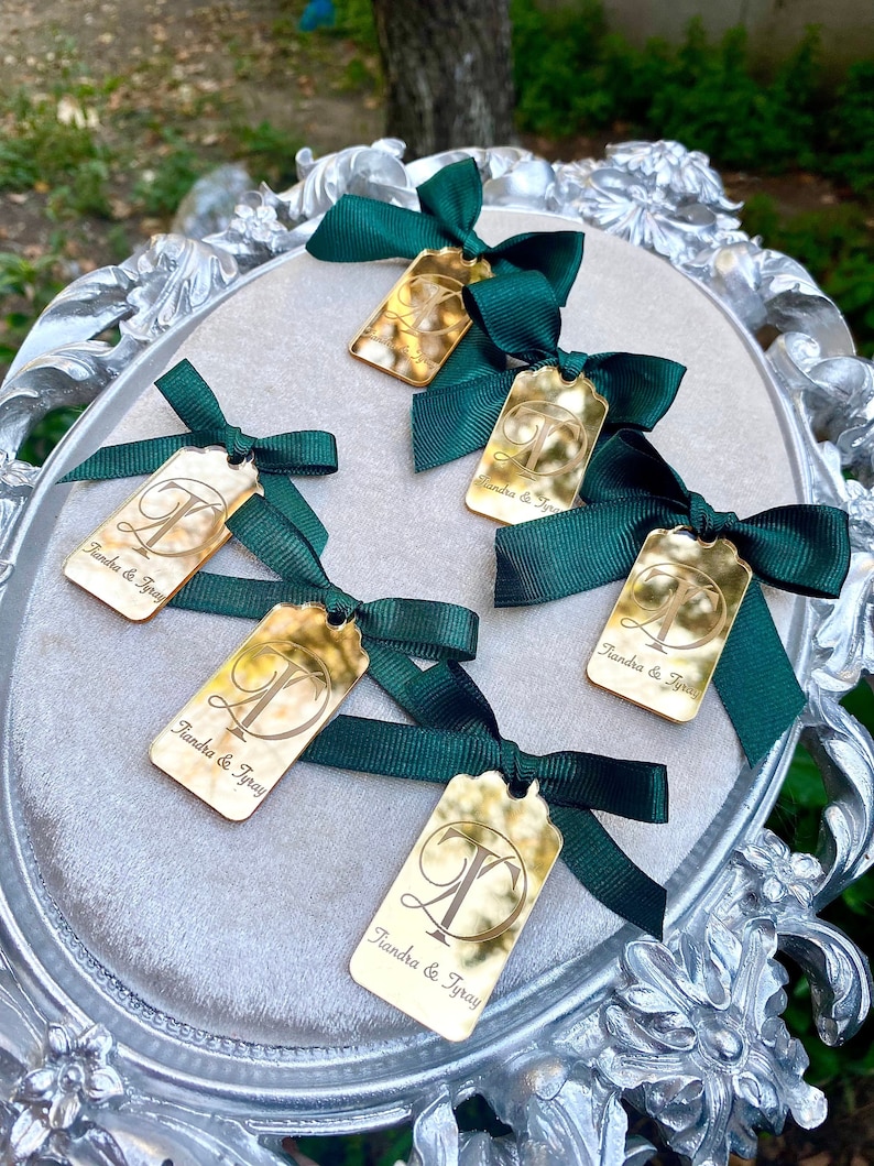 May include: Five gold mirror acrylic tags with green satin ribbon bows. Each tag has the text "Tiandra & Tyray" and a letter T in a decorative font.