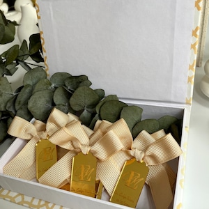 May include: A white box with a gold patterned design is open and filled with gold-colored tags. Each tag has a gold ribbon tied around it and the letter "E" engraved on it. The tags also have the text "Dr. Evelyn Marie" and a date.