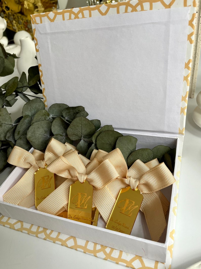 May include: A white box with a gold patterned design is open and filled with gold-colored tags. Each tag has a gold ribbon tied around it and the letter "E" engraved on it. The tags also have the text "Dr. Evelyn Marie" and a date.