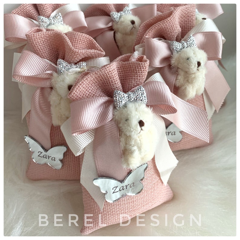 May include: Pink fabric gift bags with white teddy bears, pink bows, and silver butterfly charms with the name "Zara" on them. The bags are tied with white and pink ribbon.