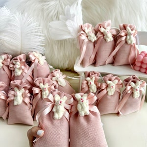 May include: Pink burlap favor bags with white bunny appliques and pink satin ribbon. The bags are arranged in a group on a white surface.