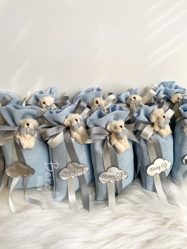Puede incluir: Bolsas de regalo azul claro con un peque&ntilde;o osito de peluche que lleva una pajarita. Cada bolsa tiene una etiqueta de nube plateada con el texto "Baby CJ" en ella.