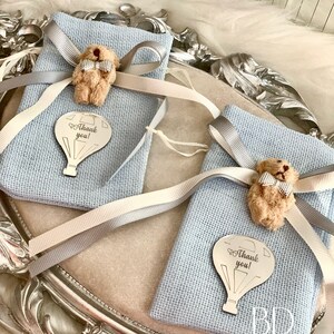 Puede incluir: Dos bolsas de regalo azules con cinta blanca y gris, cada una con un peque&ntilde;o osito de peluche con pajarita y un amuleto de globo aerost&aacute;tico plateado que dice "&iexcl;Gracias!"