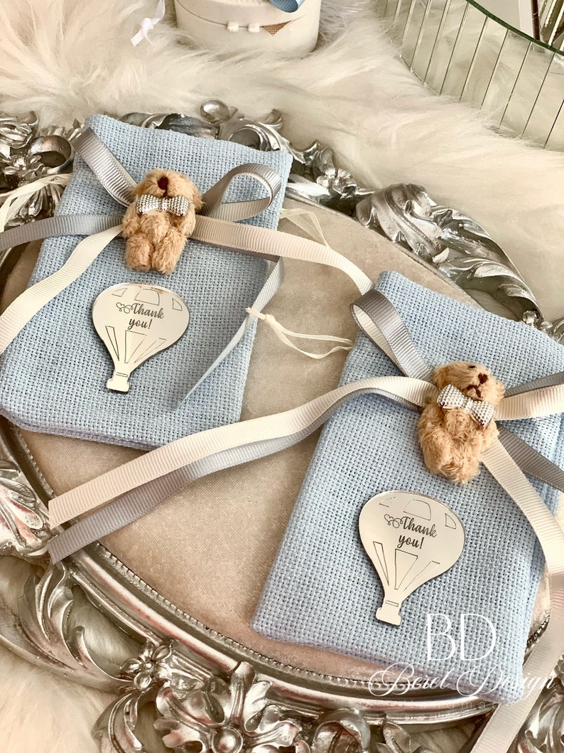 Puede incluir: Dos bolsas de regalo azules con cinta blanca y gris, cada una con un peque&ntilde;o osito de peluche con pajarita y un amuleto de globo aerost&aacute;tico plateado que dice "&iexcl;Gracias!"