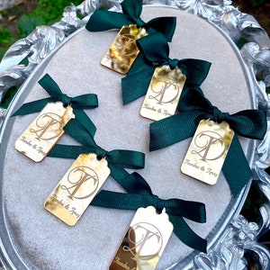 May include: Five gold-colored rectangular tags with a decorative letter "T" and the text "Tiandra & Tyray" engraved on each. Each tag is tied with a dark green ribbon bow.