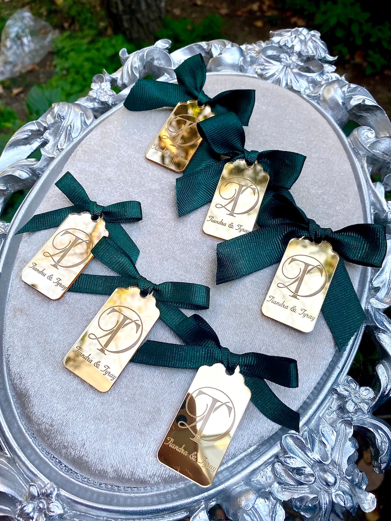 May include: Five gold-colored rectangular tags with a decorative letter "T" and the text "Tiandra & Tyray" engraved on each. Each tag is tied with a dark green ribbon bow.