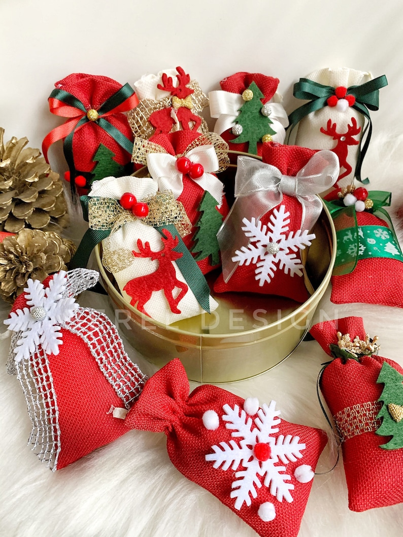 Puede incluir: Una colecci&oacute;n de peque&ntilde;as bolsas de arpillera roja y blanca con diversas decoraciones navide&ntilde;as, incluyendo copos de nieve, renos y &aacute;rboles de Navidad. Las bolsas est&aacute;n dispuestas en un recipiente de metal dorado.